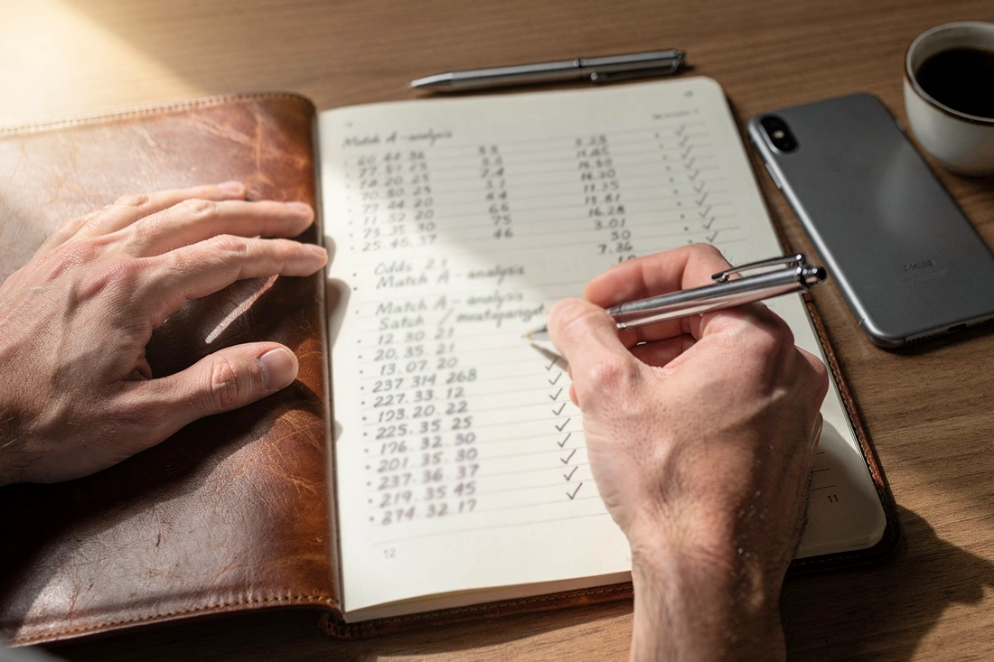 Hands writing organized betting notes in leather notebook with pen and coffee on desk