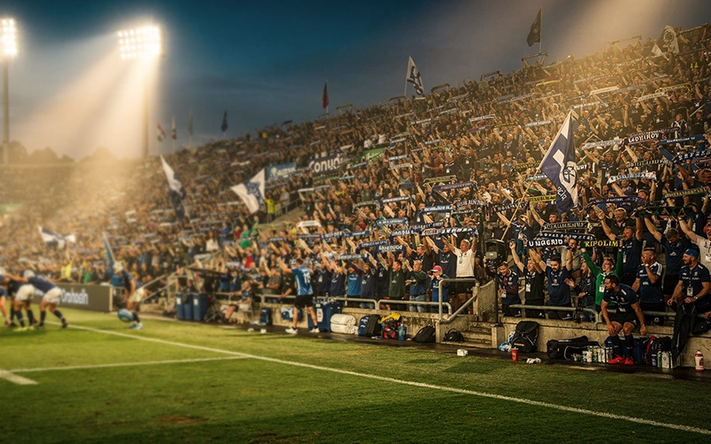 Packed rugby stadium with passionate home crowd creating intense atmosphere under floodlights