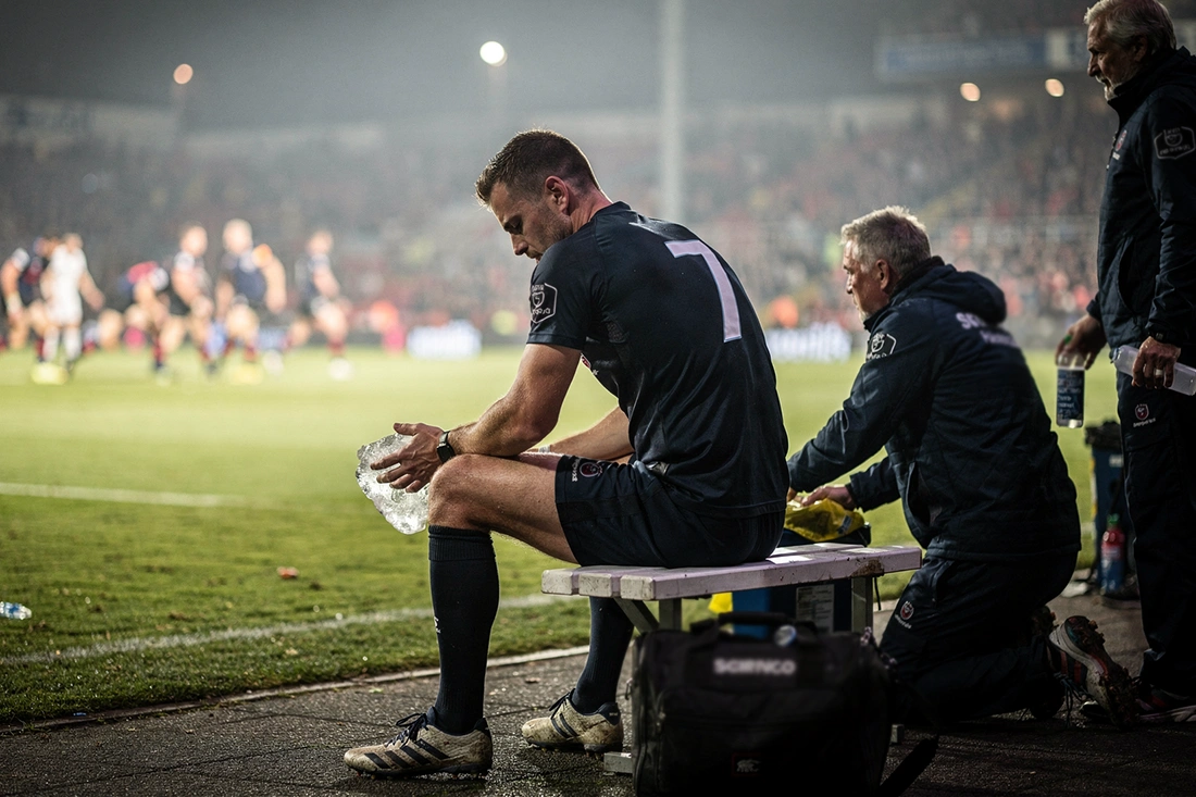 Rugby player sitting on sideline bench with ice pack receiving treatment from medical staff during match